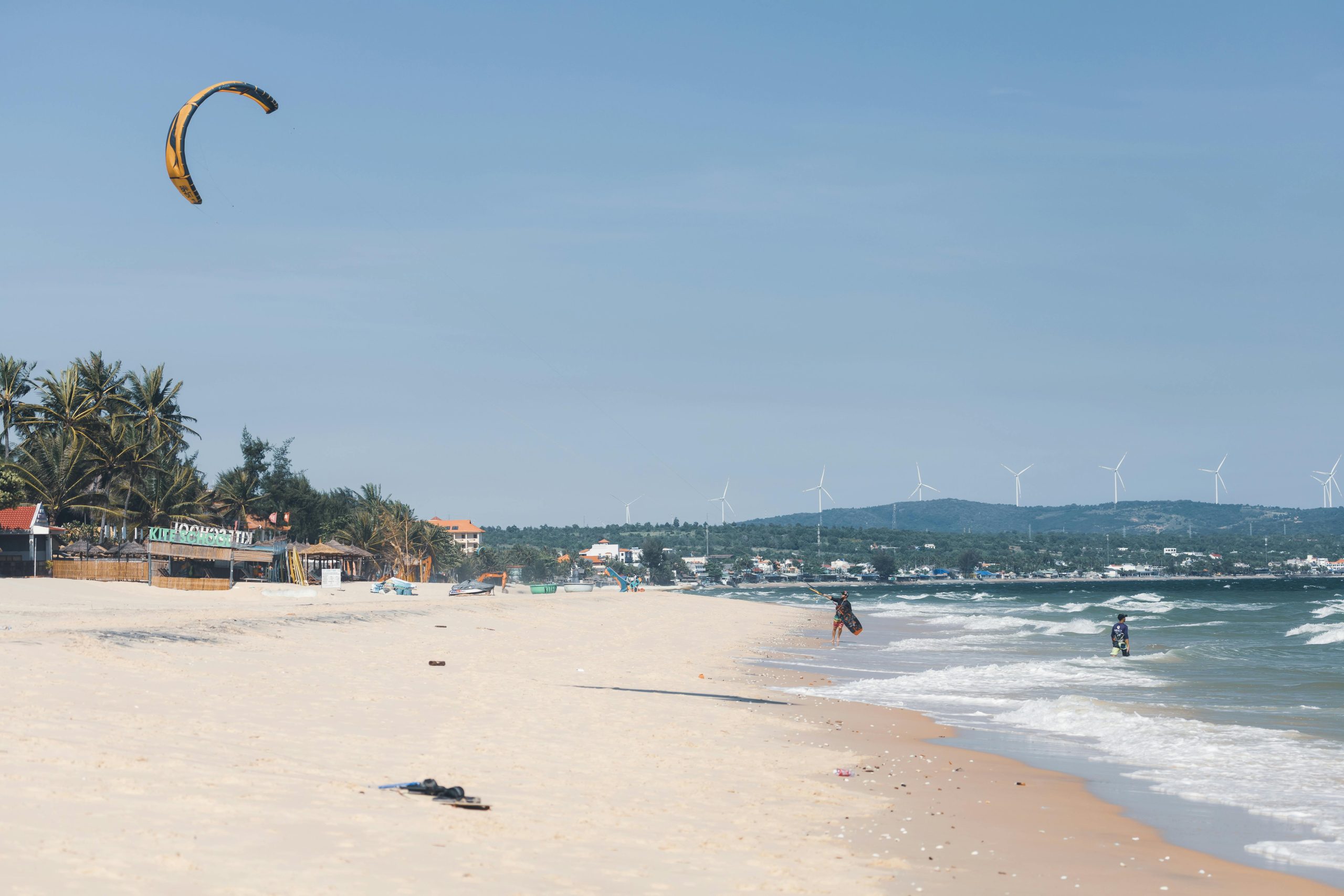 Kite surfer enjoying a sunny day on a tropical beach with wind turbines in the background.